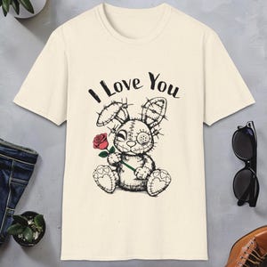 Puede incluir: Una camiseta de color crema con una ilustración en blanco y negro de un conejito cosido que sostiene una rosa roja. El texto "I Love You" está impreso sobre el conejito.