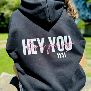May include: Black hooded sweatshirt with the text "HEY YOU This is your sign 11:11" printed on the back in white and pink. The sweatshirt is made of a soft material and has a drawstring hood.