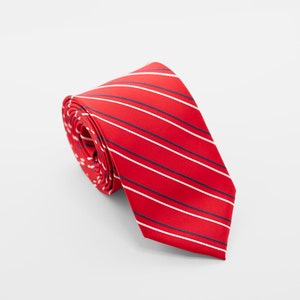May include: Rolled red tie with white and navy blue stripes. The tie is made of a smooth fabric and is displayed on a white background. The stripes are evenly spaced across the tie.