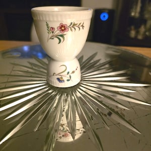 May include: A white porcelain egg cup featuring a flared rim and a tapered base. The exterior is decorated with hand-painted floral motifs in shades of pink, green, and blue, including small blossoms and delicate green leaves.