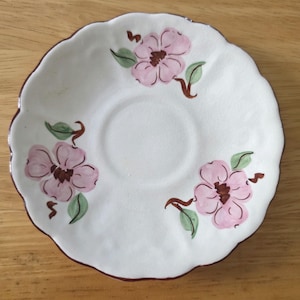 May include: A white ceramic saucer with a scalloped edge and a dark brown rim. The saucer features three hand-painted pink flowers with dark brown centers and green leaves arranged symmetrically around the central indentation of the dish.