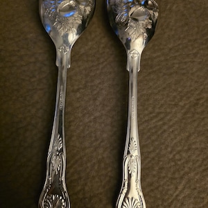 May include: Two silver-toned metal serving spoons with polished finishes. The bowls feature embossed fruit and leaf designs, while the handles display intricate scrollwork and a shell motif at the base. Each handle is engraved with text.