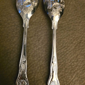 May include: Two silver-toned serving spoons featuring an embossed fruit and leaf design on the bowl. The handles have a decorative scrollwork pattern and a shell motif at the base. The text "F.B. ROGERS ISRAEL" is engraved on the handle stems.