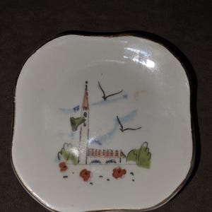 May include: A small, square-shaped ceramic dish with rounded corners and a gold-trimmed edge. The center features a hand-painted illustration of a tall church tower, a building with a red roof, green bushes, orange flowers, and two black birds.