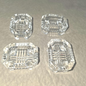 May include: A set of four clear glass rectangular dishes featuring a faceted, geometric pattern on the exterior and a grid-like embossed design on the interior base. Each dish has small, raised feet at the corners and a thick, polished rim.