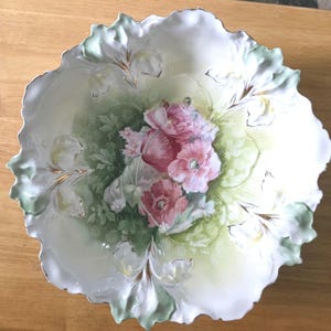 May include: A round porcelain bowl with a scalloped, ruffled edge featuring a central floral motif of pink poppies surrounded by soft green foliage. The white background transitions into pale green washes, accented with gold trim along the rim.