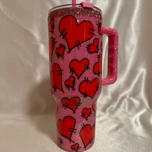 May include: A pink glitter tumbler with a handle, featuring red stitched hearts. The tumbler includes a pink straw and a glittery rim. The design is a fun, Valentine's Day theme.