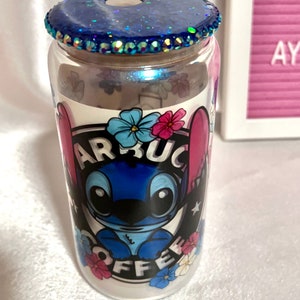 Op de afbeelding: Een doorzichtige glazen beker met een blauw glitterdeksel en een blauw cartoonfiguur met de tekst "Starbucks Coffee" aan de zijkant.