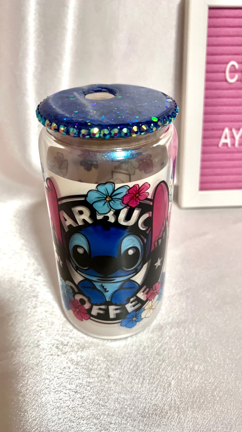 Op de afbeelding: Een doorzichtige glazen beker met een blauw glitterdeksel en een blauw cartoonfiguur met de tekst "Starbucks Coffee" aan de zijkant.