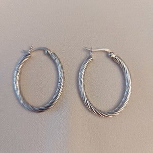 May include: A pair of silver hoop earrings. Each earring features a twisted rope design around the oval shape. The earrings are displayed against a neutral background.