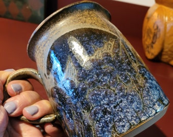 May include: A blue and brown ceramic mug with a speckled glaze. The mug has a handle and is held in a hand.