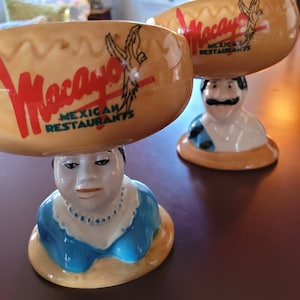 Pode incluir: Duas tigelas de cerâmica com base marrom e amarela e um logotipo vermelho e branco que diz "Mocano Mexican Restaurants". As tigelas têm a forma de um sombrero e são coroadas com um busto de cerâmica de uma pessoa que usa uma camisa azul.