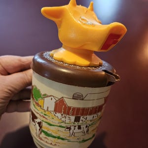 May include: A vintage ceramic creamer with a brown and white farmhouse scene featuring a red barn and cows. The lid is a yellow plastic cow head with a red nose.