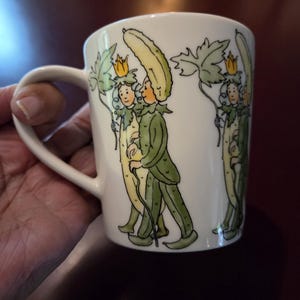 May include: White ceramic mug with a whimsical illustration of figures dressed as cucumbers, holding leaves and wearing crowns. The figures are in green, yellow, and white. The mug has a curved handle.