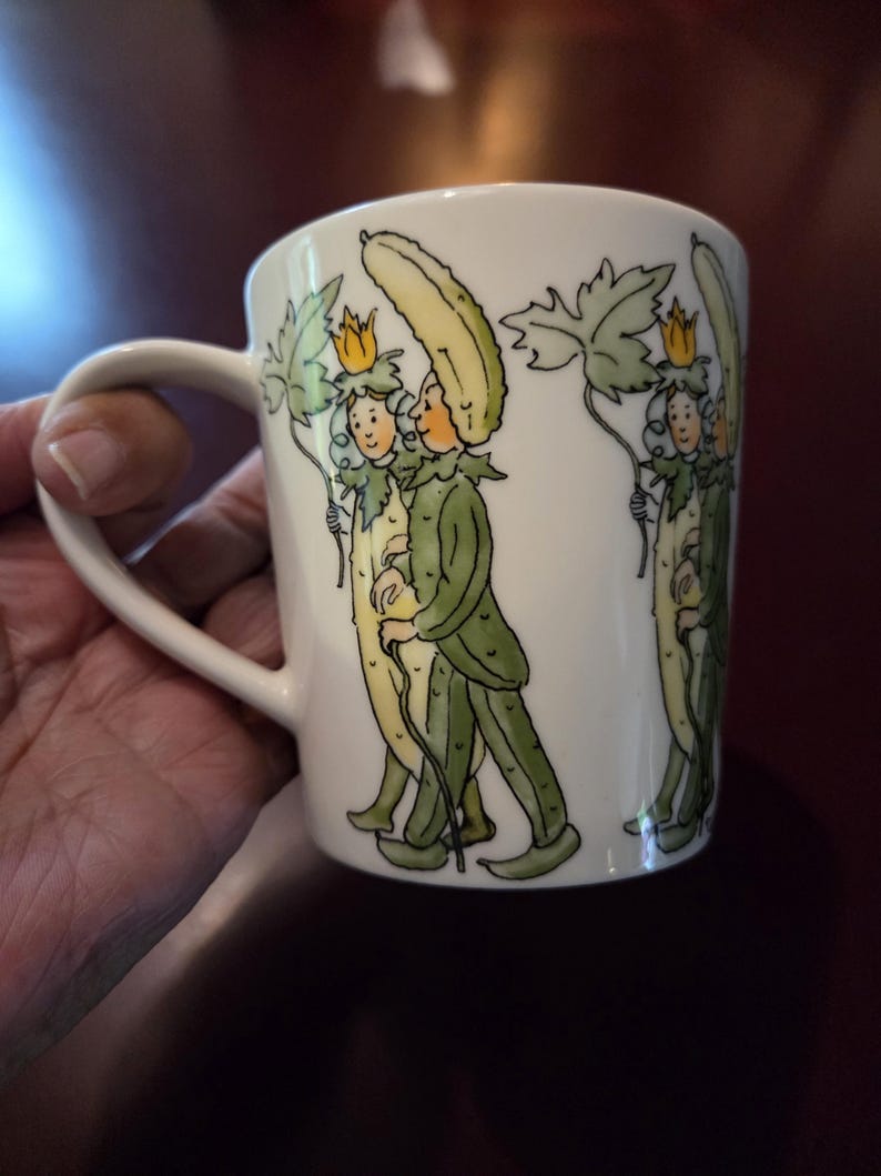May include: White ceramic mug with a whimsical illustration of figures dressed as cucumbers, holding leaves and wearing crowns. The figures are in green, yellow, and white. The mug has a curved handle.