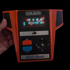 May include: Vintage Coleco Alien Attack handheld game. The orange and black game features a digital display showing the game's graphics. It has red and blue buttons labeled "FIRE" and "MOVE", and a directional pad. The game's title is printed at the bottom.