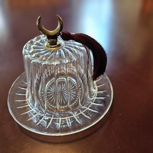 May include: A clear glass dome with a decorative pattern and a crescent moon finial. The dome sits on a matching clear glass base with a starburst pattern. A burgundy tassel hangs from the finial.