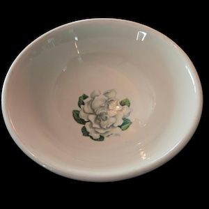 May include: A white ceramic bowl with a floral design. The bowl features a white gardenia flower with green leaves in the center. The bowl has a smooth, glossy finish and a rounded shape. The background is black.