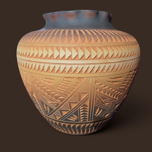 May include: A terracotta-colored ceramic vase with a wide, rounded body and a ruffled rim. The surface features intricate, carved geometric patterns in dark gray and natural clay tones, including repeating triangles and linear motifs.