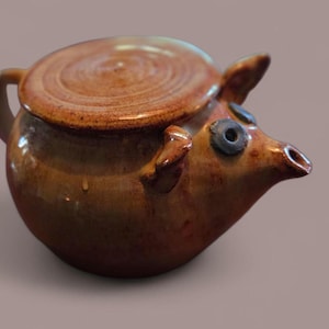 May include: A ceramic teapot with a pig design. The teapot is brown with a lid and a handle. The pig's face has blue eyes and a snout. The teapot is a unique and decorative item.