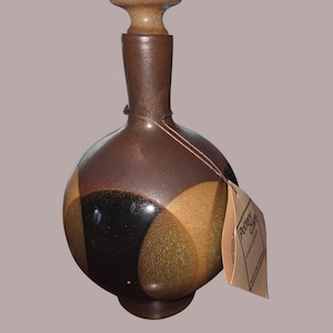 May include: A brown ceramic decanter with a round body and a stopper. The decanter has a dark brown neck and a light brown stopper. The body features a geometric design in shades of brown, black, and tan. A tag is attached to the decanter.