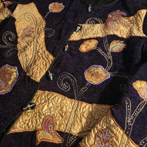 May include: A dark purple textured jacket featuring gold-coloured fabric panels and intricate floral embroidery in shades of orange, yellow, and purple. The garment includes a front button closure with dark, metallic-toned toggle-style buttons.