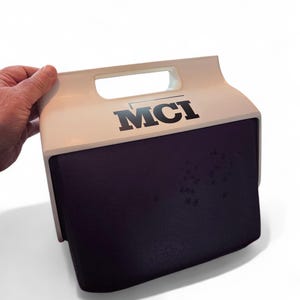 Puede incluir: Una nevera portátil blanca y morada con asa. La nevera tiene las letras "MCI" impresas en negro en la parte superior. La nevera es sostenida por una mano.
