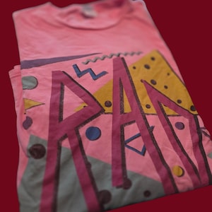 Può includere: Una maglietta rosa con un design geometrico nei toni del viola, giallo e blu. Il design presenta la parola "RA" in grandi lettere stilizzate, insieme a varie forme e motivi. La maglietta è piegata e posta su uno sfondo rosso scuro.