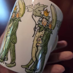 May include: White ceramic mug with a whimsical illustration of figures dressed as cucumbers, with green and yellow details. The figures wear crowns and hold a leaf. The mug has a handle and is held by a hand.