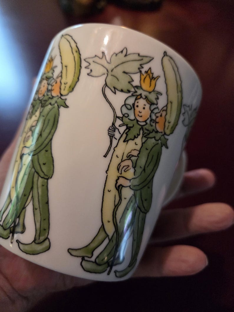 May include: White ceramic mug with a whimsical illustration of figures dressed as cucumbers, with green and yellow details. The figures wear crowns and hold a leaf. The mug has a handle and is held by a hand.