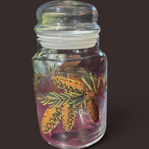 May include: A clear glass canister with a removable lid featuring a painted design of brown pinecones and green pine needles on the exterior. The cylindrical container has a smooth surface and a translucent appearance, showcasing the botanical art.