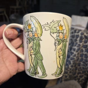 May include: White ceramic mug with a whimsical illustration of figures in green suits and yellow hats. The figures are holding long, yellow objects and walking. The mug has a curved handle.