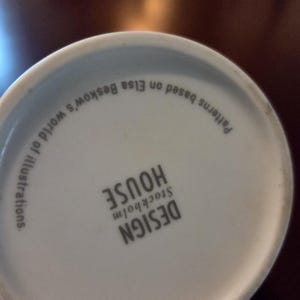 May include: White ceramic mug base with the text "DESIGN HOUSE STOCKHOLM" and "Patterns based on Elsa Beskow's world of illustrations." The text is in black. The mug is on a dark brown surface.