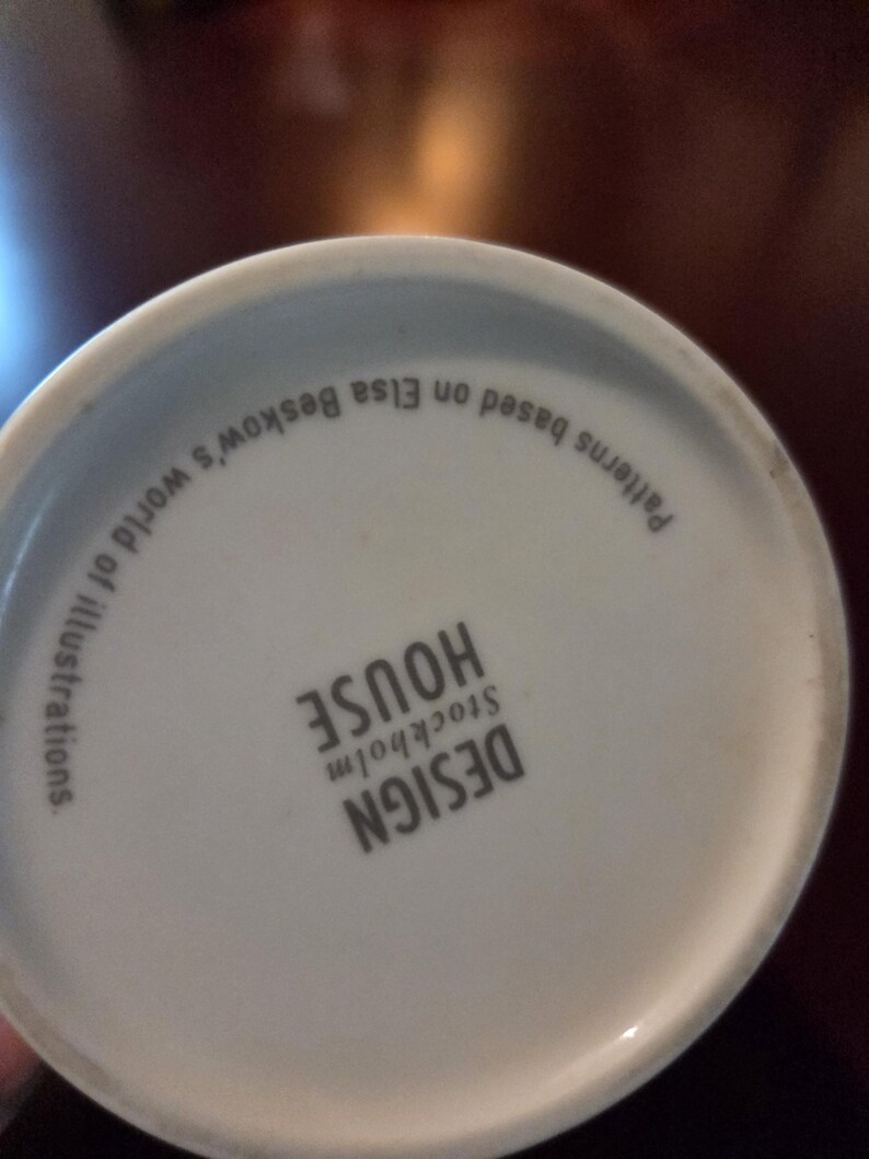May include: White ceramic mug base with the text "DESIGN HOUSE STOCKHOLM" and "Patterns based on Elsa Beskow's world of illustrations." The text is in black. The mug is on a dark brown surface.