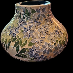 Puede incluir: Un jarrón decorativo con forma bulbosa, con un patrón floral en tonos de azul, verde y crema. Las flores y hojas están pintadas sobre la superficie, creando una apariencia texturizada. El jarrón tiene un interior azul oscuro.