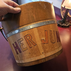 Puede incluir: Un cubo de madera con una banda de metal alrededor de la parte superior. El cubo está pintado con las palabras "HER JUNK" en letras rojas y naranjas.
