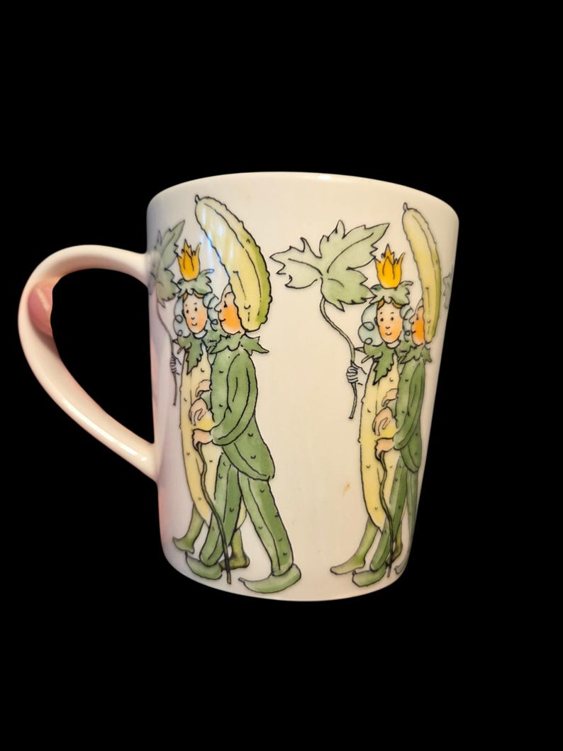 May include: White ceramic mug with a repeating illustration of figures in green and yellow cucumber costumes, carrying leaves and walking with canes. The design wraps around the mug, which has a curved handle on the left side.