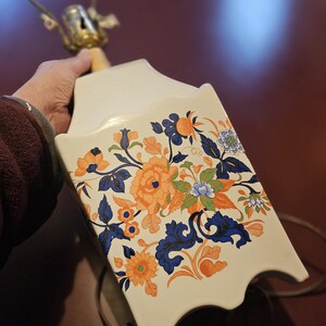 May include: White ceramic lamp base with a floral design in shades of blue, orange, and green. The base has a scalloped edge and a gold-colored metal top.