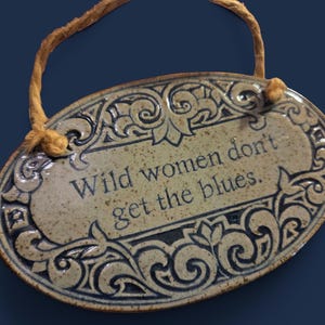 May include: An oval-shaped ceramic wall plaque with a speckled beige finish and dark blue decorative scrollwork borders. The text "Wild women don't get the blues." is centered on the plaque, which hangs from a tan-colored fiber cord.