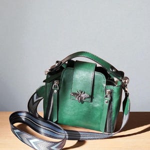 May include: Green leather handbag with a silver bee emblem on the front flap. The bag has a top handle, two zippered compartments, and a patterned shoulder strap. The bag is set against a neutral background.