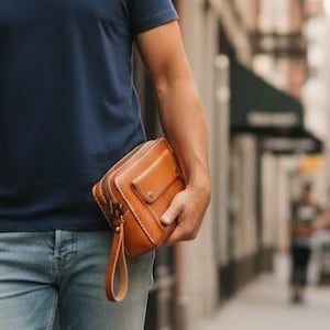 May include: A navy blue crew neck t-shirt, light blue denim jeans, and white canvas sneakers. A tan leather rectangular clutch bag with a wrist strap is held in one hand. The outfit consists of a simple short-sleeved shirt and slim-fit jeans.