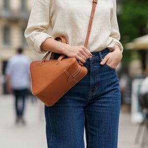 Peut inclure: Une personne porte une chemise crème à manches longues et un jean bleu foncé. Elle porte un sac à main en cuir marron avec une bandoulière. En arrière-plan, une scène de rue européenne.