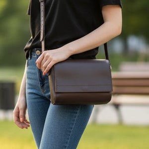 May include: A brown leather crossbody bag with a flap closure and adjustable strap. The bag is worn over a black t-shirt and blue jeans. White sneakers complete the outfit. The background is a park setting with green grass and trees.