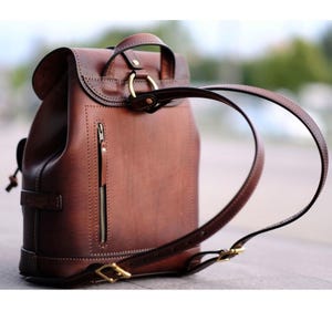 Peut inclure: Un sac à dos en cuir marron avec une fermeture à rabat et une poignée supérieure. Le sac à dos a une poche verticale zippée sur le devant et des bretelles réglables. La quincaillerie est de couleur dorée.