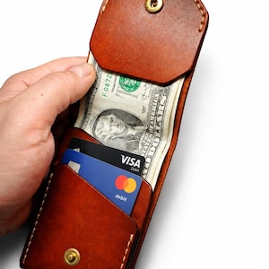 Puede incluir: Una cartera de cuero marrón con cierre de botón a presión, que contiene un billete de dólar estadounidense y dos tarjetas de crédito. La cartera está abierta, revelando su contenido. El cuero tiene un grano natural y un tono marrón rojizo cálido.