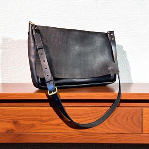 May include: A black leather messenger bag with a long adjustable strap and a buckle closure. The bag has a flap closure and is sitting on a wooden surface. The bag's hardware is gold-colored.