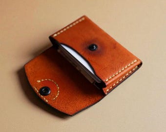 May include: Open, brown leather card holder with a snap closure. The card holder is open, revealing white cards inside. The edges are stitched with a contrasting thread. The background is a solid tan color.