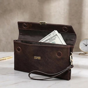 Puede incluir: Un bolso de mano tipo sobre en cuero marrón oscuro con correa de muñeca, que presenta un cierre metálico rectangular en tono latón, costuras visibles y una solapa abierta que revela un fajo de billetes estadounidenses en su interior.