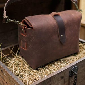 May include: A brown leather messenger bag featuring a fold-over flap with a dark brown leather strap closure, brass-toned metal hardware, and an adjustable shoulder strap attached with swivel clips on the sides of the bag.
