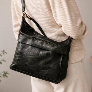 May include: A black leather shoulder bag with a flap closure and a long strap. The bag has a distressed finish and a gold-colored metal ring on the strap. The bag is worn over the shoulder.